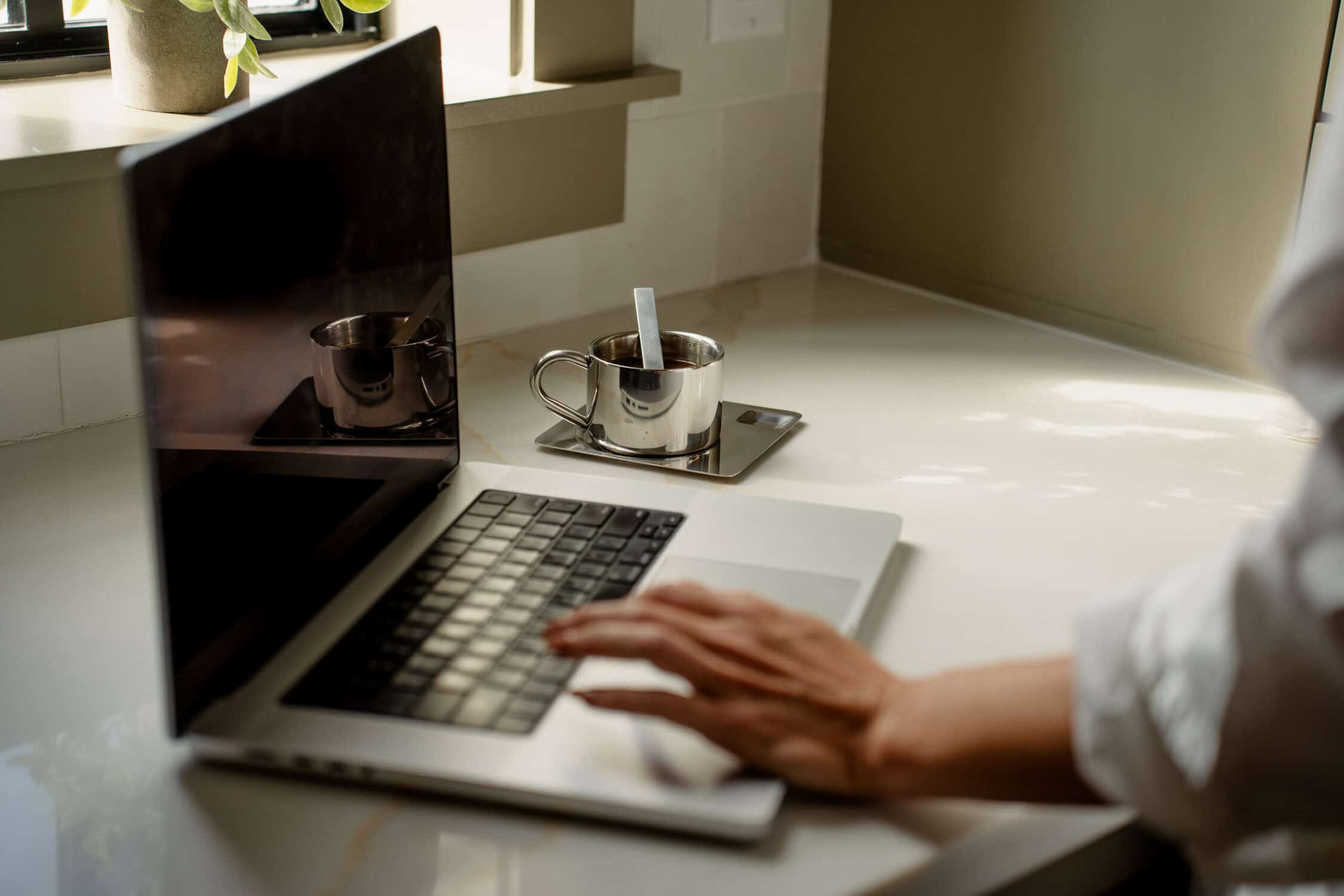 De hand van een persoon rust op het toetsenbord van een laptop op een licht aanrecht. Terwijl het zonlicht naar binnen stroomt, overdenken ze verschillende soorten websites, met een metalen kopje in de buurt en groene bladeren in de hoek. Op het scherm van de laptop is een reflectie te zien.