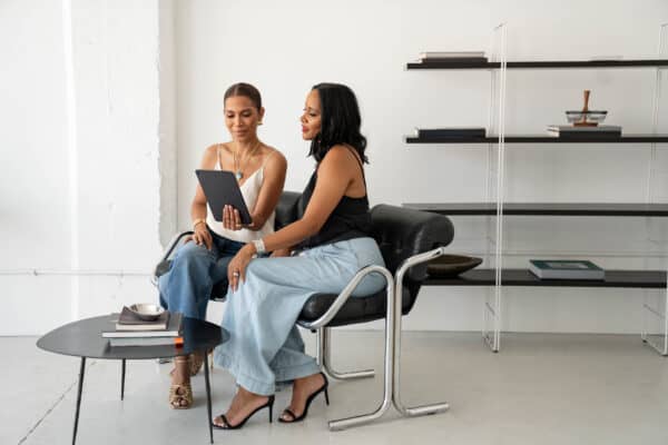 Twee vrouwen zitten samen in een modern, minimalistisch kantoor. Ze zijn bezig met het opbouwen van een gemeenschap en bespreken ideeën via een tablet. Een ronde tafel met boeken en een lege plank benadrukken de schone omgeving. Beiden zijn gekleed in lichte broeken en mouwloze topjes.