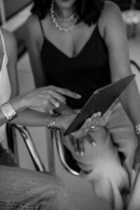 Twee vrouwen zitten naast elkaar, van bovenaf gezien in zwart-wit. De ene in een donker hemdje en halsketting houdt een tablet vast, terwijl de andere, met een lichte tanktop en armbanden, naar het scherm wijst. Hun gezichten zijn gedeeltelijk buiten beeld.