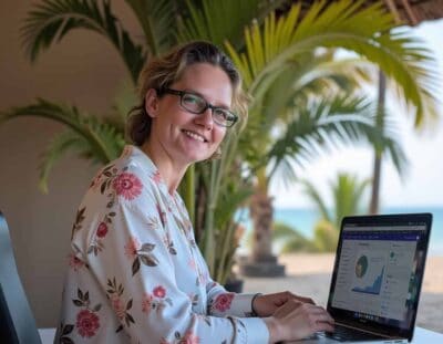 Een vrouw met een bril en een gebloemde blouse glimlacht naar de camera terwijl ze buiten op haar laptop aan website onderhoud werkt. Palmbomen en een strand met blauwe lucht en oceaan zijn zichtbaar op de achtergrond, samen met een grafiek op haar scherm.