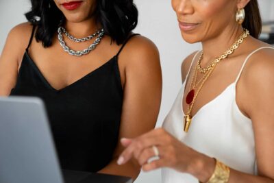 Twee vrouwen met mouwloze topjes en gelaagde gouden en zilveren sieraden werken samen aan een laptop. Als merkambassadeurs zijn ze gefocust op het opbouwen van een gemeenschap, waarbij de een naar het scherm wijst terwijl ze samen strategieën bedenken.