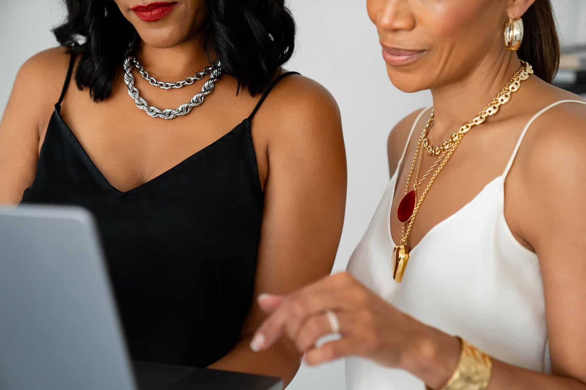 Twee vrouwen met mouwloze topjes en gelaagde gouden en zilveren sieraden werken samen aan een laptop. Als merkambassadeurs zijn ze gefocust op het opbouwen van een gemeenschap, waarbij de een naar het scherm wijst terwijl ze samen strategieën bedenken.