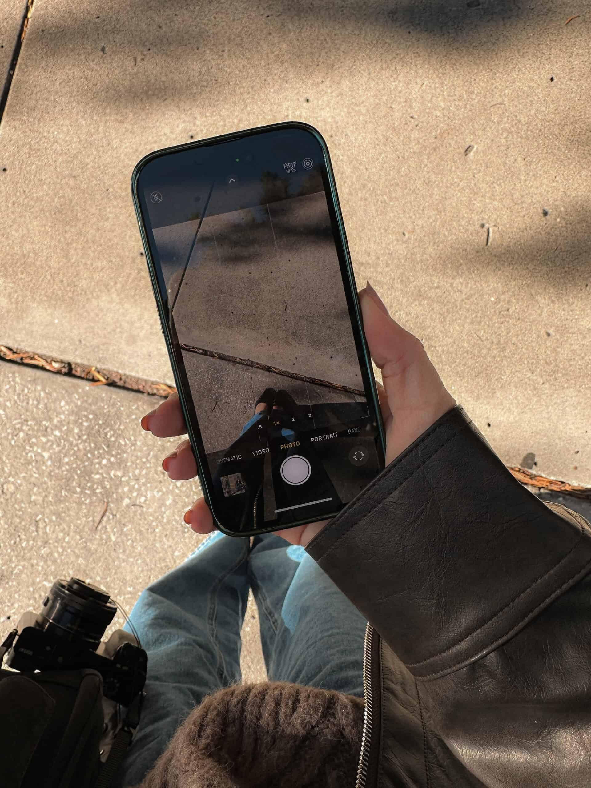 Een persoon met een donker leren jack en een blauwe spijkerbroek houdt een smartphone vast en neemt een foto van zijn schaduw op een zonovergoten stoep. Het telefoonscherm toont de camera-interface met hun schoenen en het trottoir zichtbaar. Een camera hangt aan hun zijde.