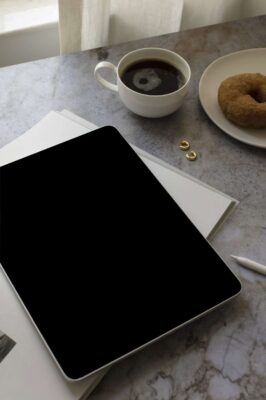 Een tablet met een leeg zwart scherm rust op een gesloten notitieblok op een marmeren tafel. Vlakbij zijn tekenen van klantinteractie - twee gouden ringen en koffie - vermengd met merkbeleving in het uitnodigende licht dat door witte gordijnen naar binnen schijnt.