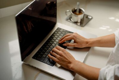 Een persoon typt op het toetsenbord van een laptop aan een bureau, met een kop koffie en een lepel in de buurt.