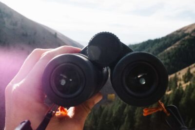 Een hand houdt een zwarte verrekijker omhoog, met bergen en bomen op de achtergrond, vaag afgebeeld tegen een gedeeltelijk bewolkte hemel.