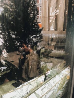 Een persoon zit op een bankje in een café en drinkt uit een kopje. Hij kijkt door een reflecterend glazen raam, en in de weerspiegeling zijn ook bomen en gebouwen zichtbaar.