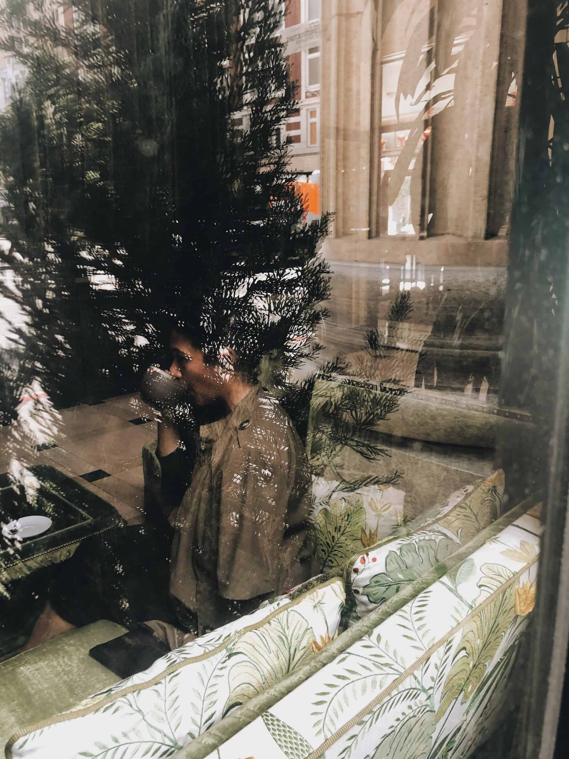 Een persoon zit op een bankje in een café en drinkt uit een kopje. Hij kijkt door een reflecterend glazen raam, en in de weerspiegeling zijn ook bomen en gebouwen zichtbaar.